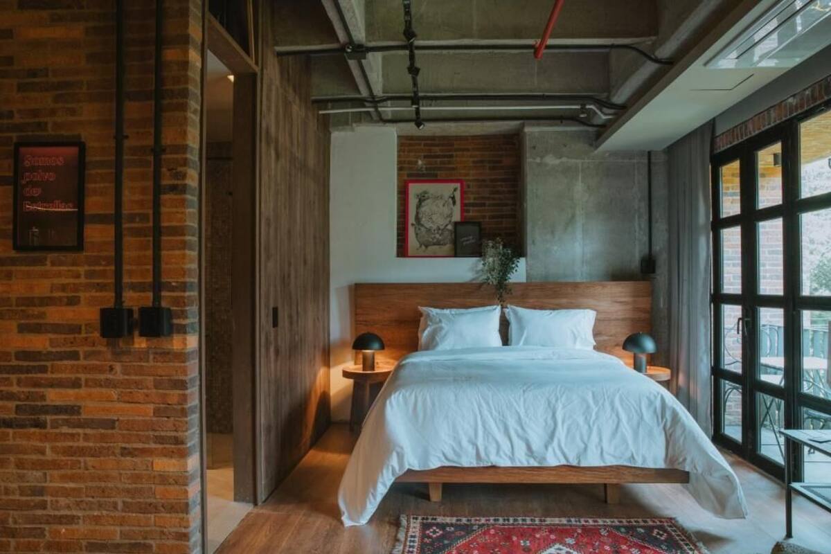 Un dormitorio con cama de madera, alfombra roja estampada, pared de ladrillo y un ventanal grande.