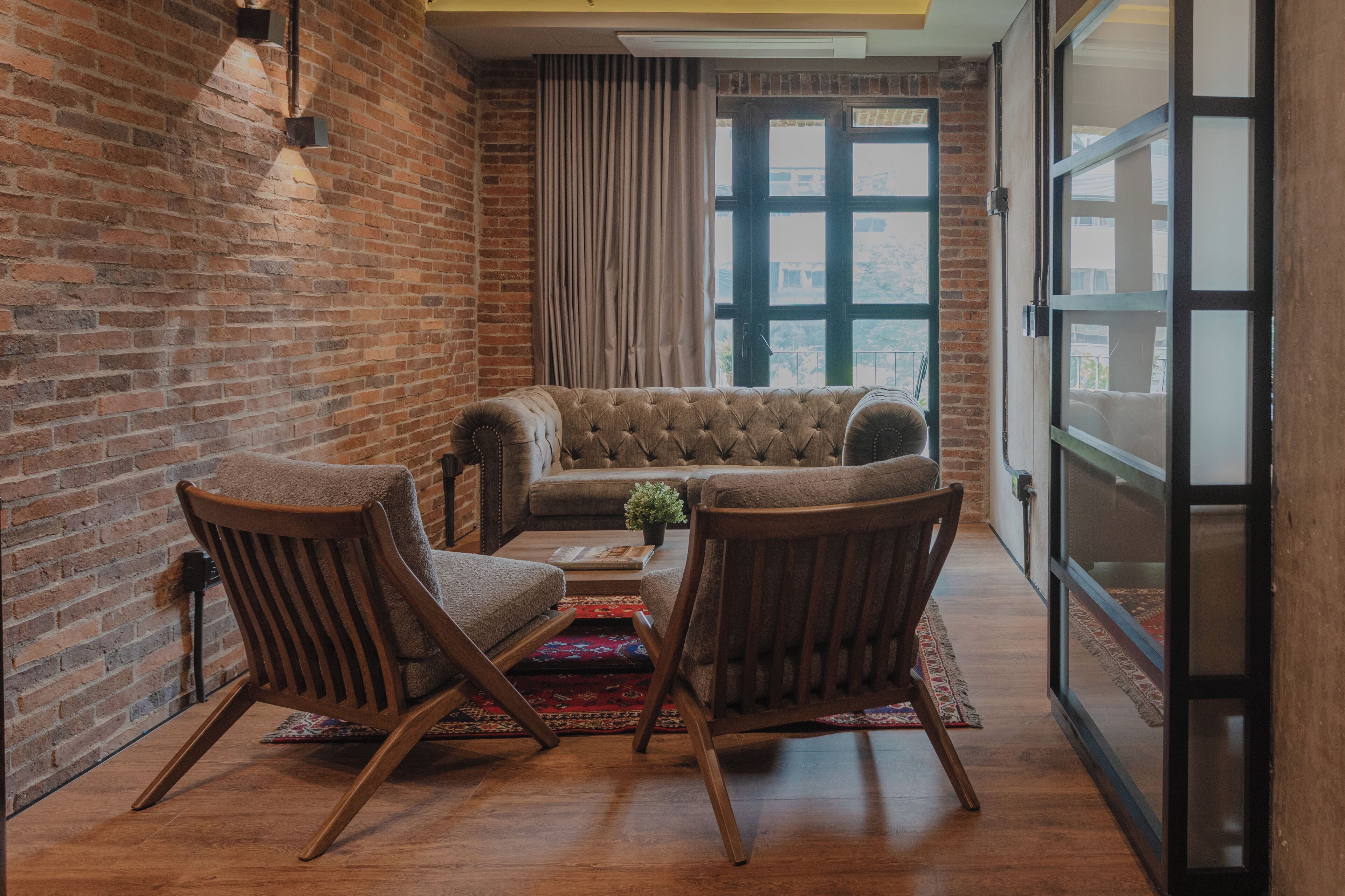A cozy room with a brick wall, a tufted sofa, two armchairs, a small table with a plant, and a view through a large window.