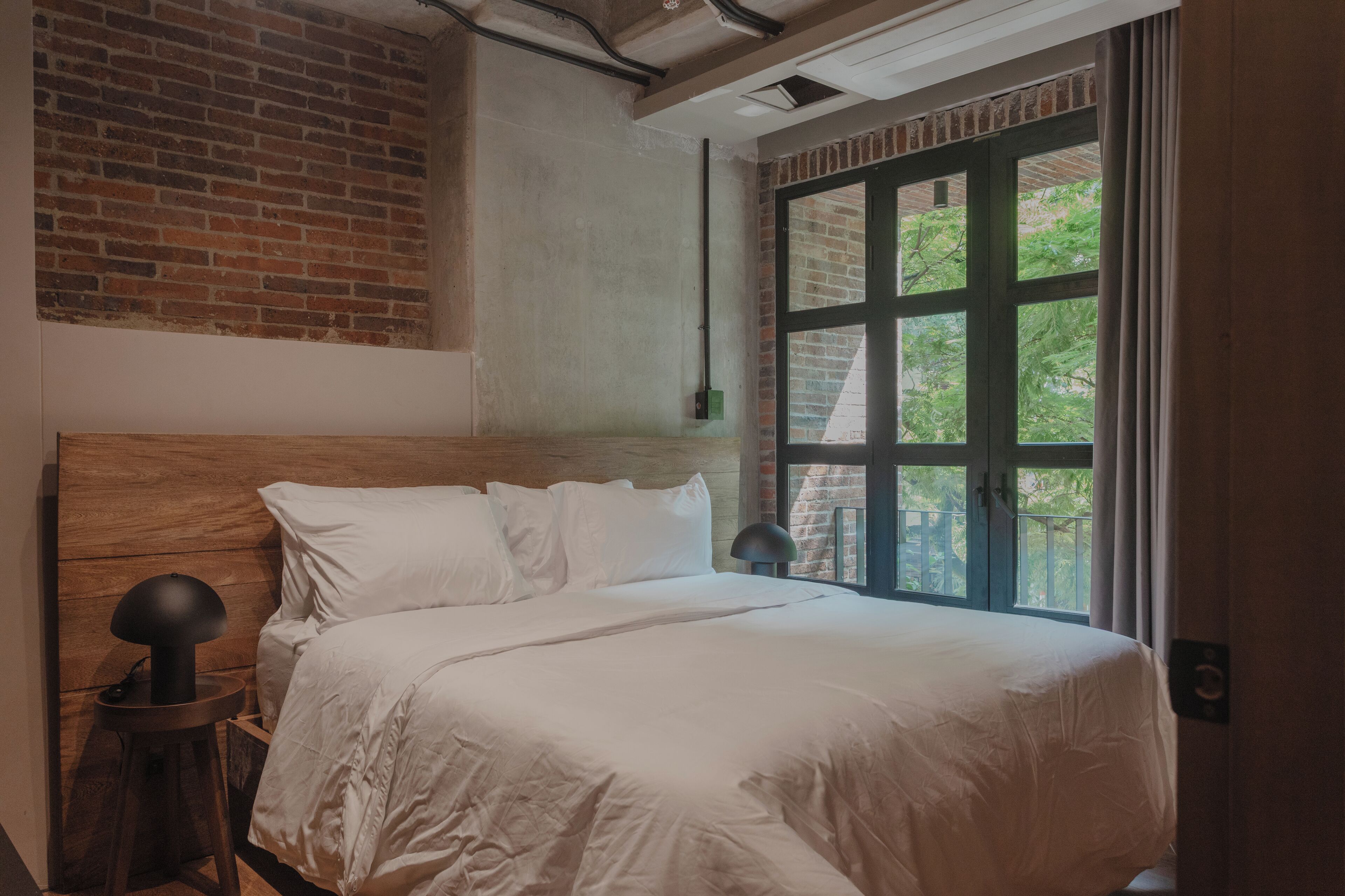 A bedroom with a wooden headboard, a large window with a view of greenery, and a modern lamp on a side table.
