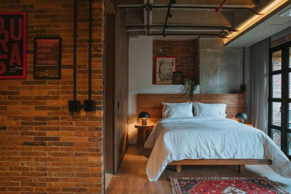 A bedroom with a brick wall, a wooden bed, a nightstand, a lamp, and a framed picture.