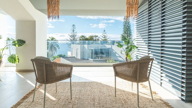 Una habitación moderna con dos sillas de mimbre, un ventanal con vistas a un edificio y palmeras, y una alfombra con textura.