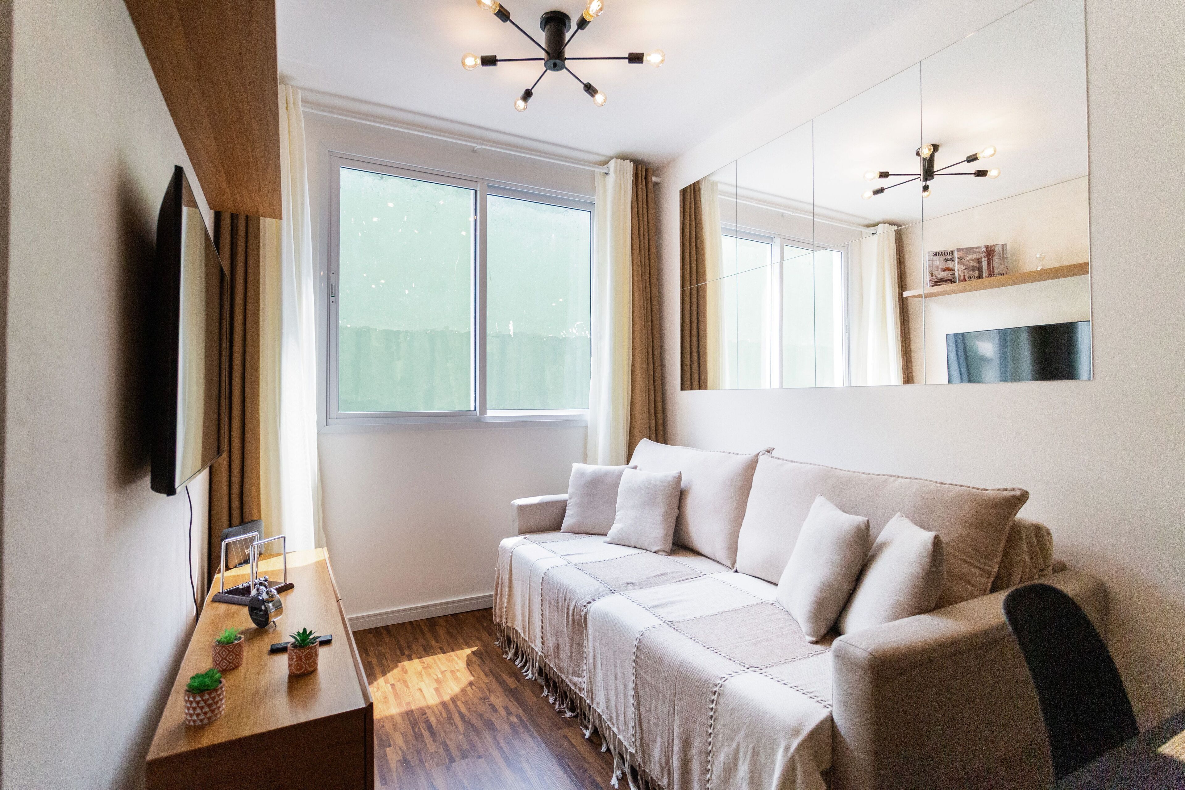 A modern living room with a sofa, a coffee table with decorative items, a TV mounted on the wall, and a large window with curtains.