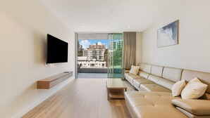 Una sala de estar moderna con un televisor de pantalla plana, un sofá color beige y un ventanal de vidrio que da a un balcón con vista a la ciudad.