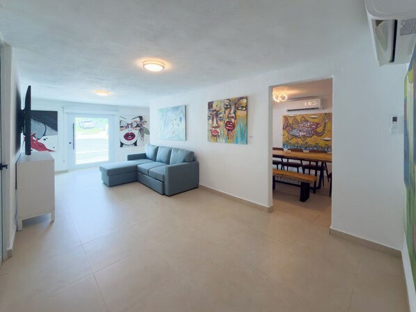 A spacious living room with a grey sectional sofa, a dining area with a wooden table and benches, and various artworks on the walls.