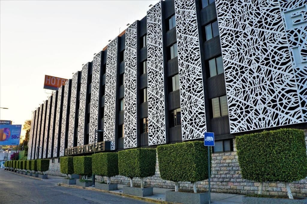 A modern hotel facade with intricate lattice patterns, a prominent 'HOTEL PRINCIPES' sign, and neatly trimmed hedges along the sidewalk.