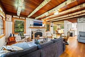 A cozy cabin interior with a fireplace, wooden ceiling beams, a television, and a dining area.