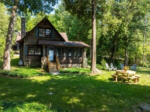 A log cabin with a stone chimney, a wooden porch, and a picnic table in a grassy yard surrounded by trees.