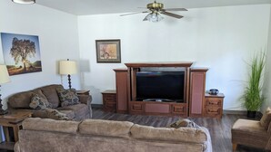 Uma sala de estar com uma grande TV de tela plana, aparador de madeira, dois sofás, uma mesa de centro, um abajur e um ventilador de teto.