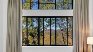A bedroom with a large window featuring a metal bar, beige curtains, and a view of trees and hills. - This is The Life - stunning mountain and lake view (Glenville)