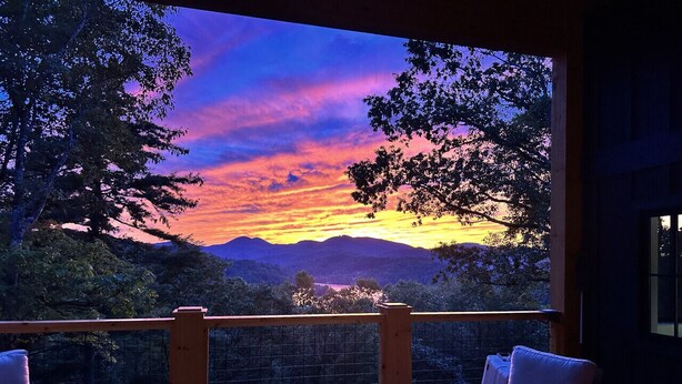 A wooden deck with two chairs and a table, overlooking a mountainous landscape during sunset. - This is The Life - stunning mountain and lake view (Glenville)