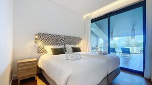 A modern bedroom with a large bed, a nightstand, and a view of the outdoors through a sliding glass door. - Acqua Beach by Atlantic Holiday (Funchal)
