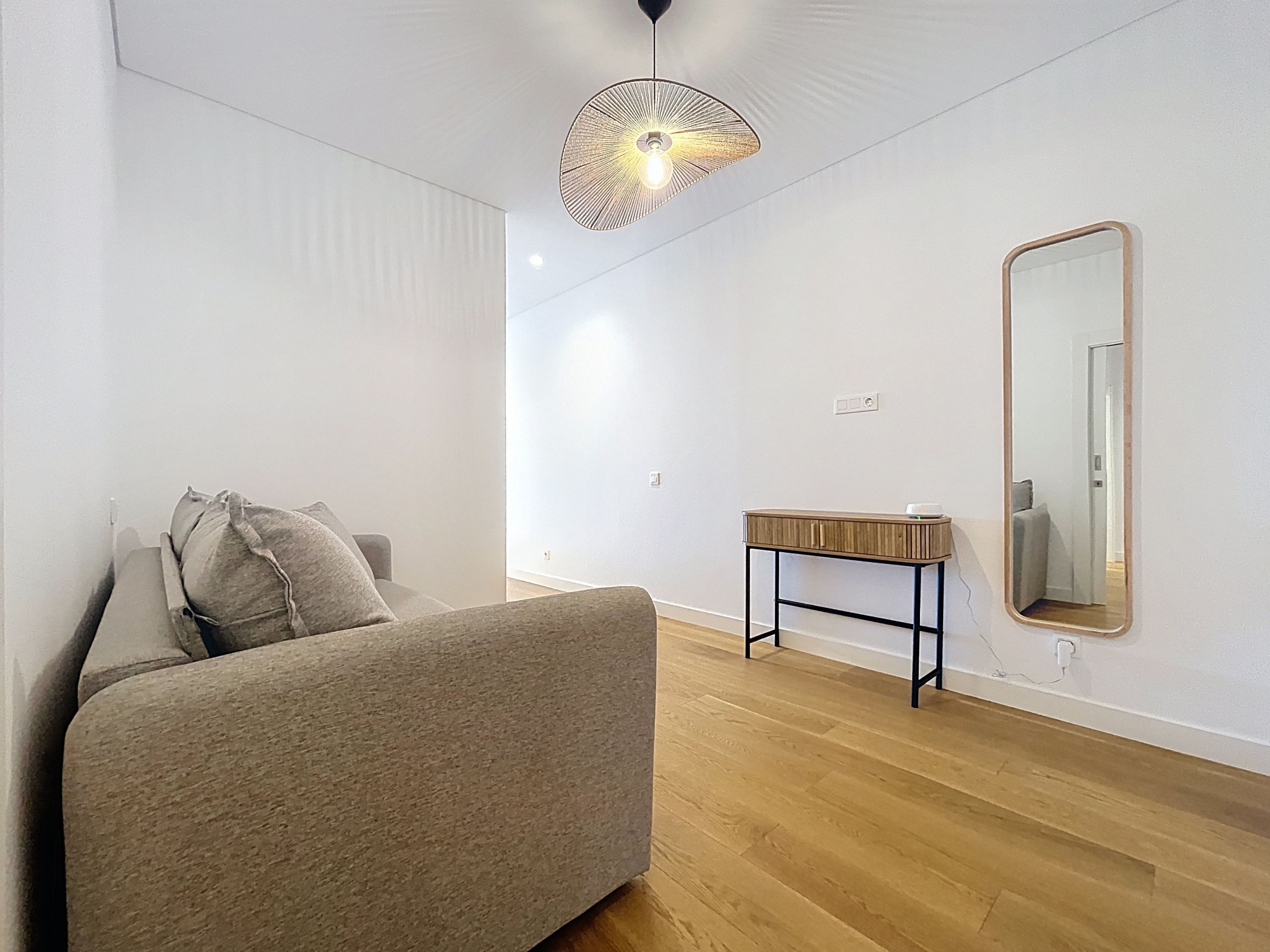 A modern, minimalist bedroom with a grey sofa, a wooden side table, a large mirror, and a unique ceiling light fixture.