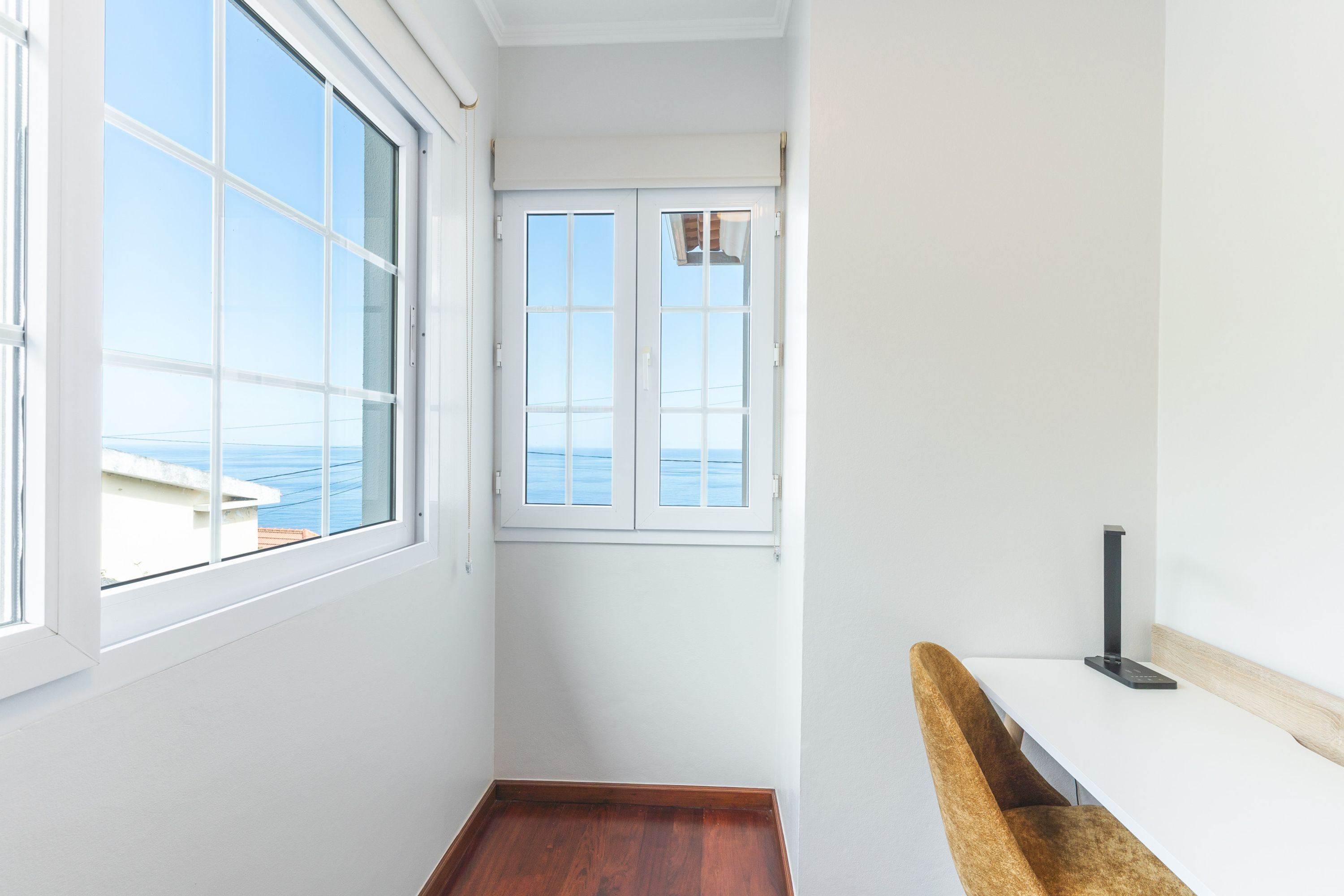 A narrow hallway with a wooden floor, a white wall, and a window with a view of the sea.