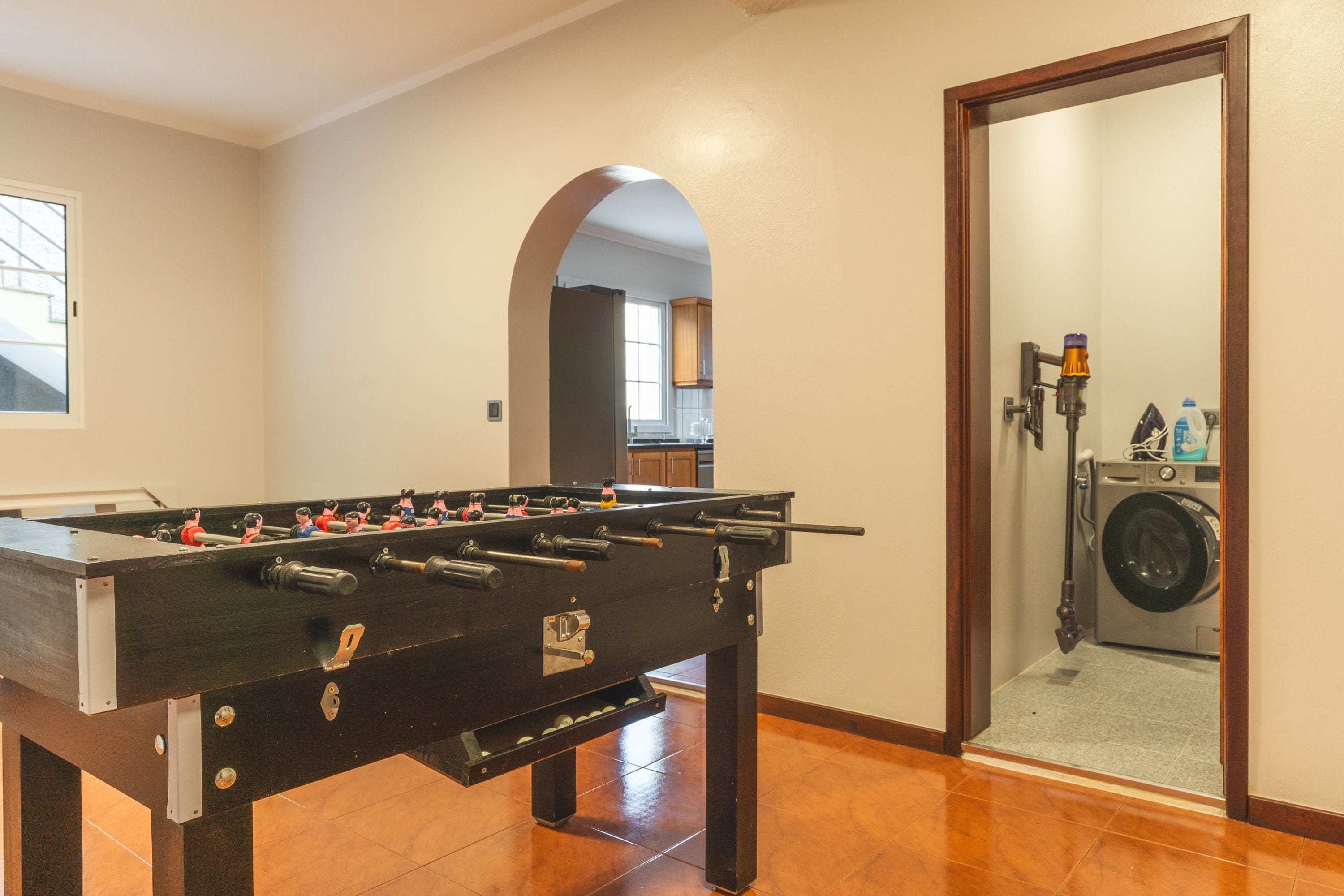 A room with a foosball table, a washing machine, and a door leading to a kitchen area.