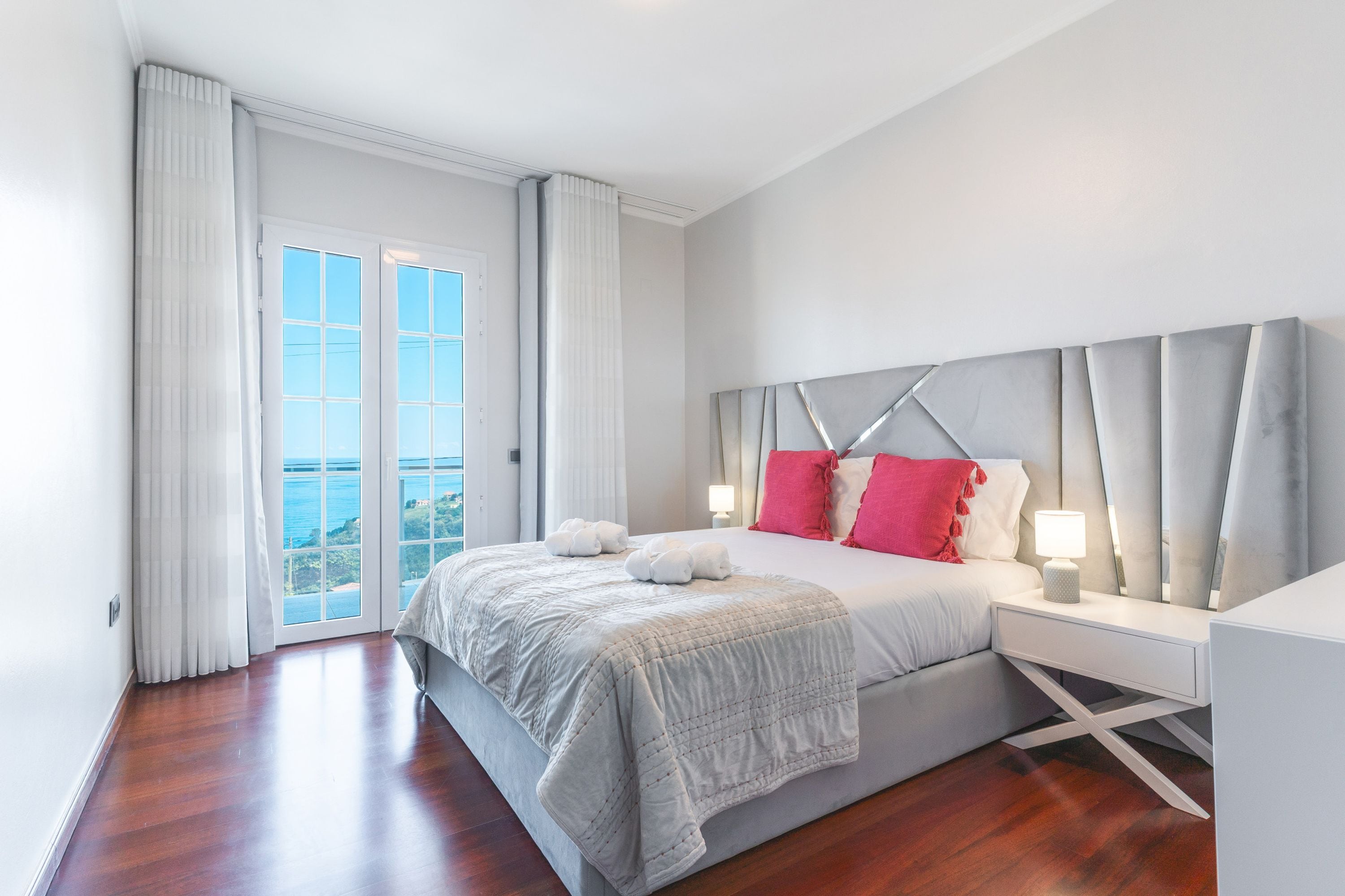 A modern bedroom with a large bed, a nightstand, and a view of the ocean through a sliding glass door.
