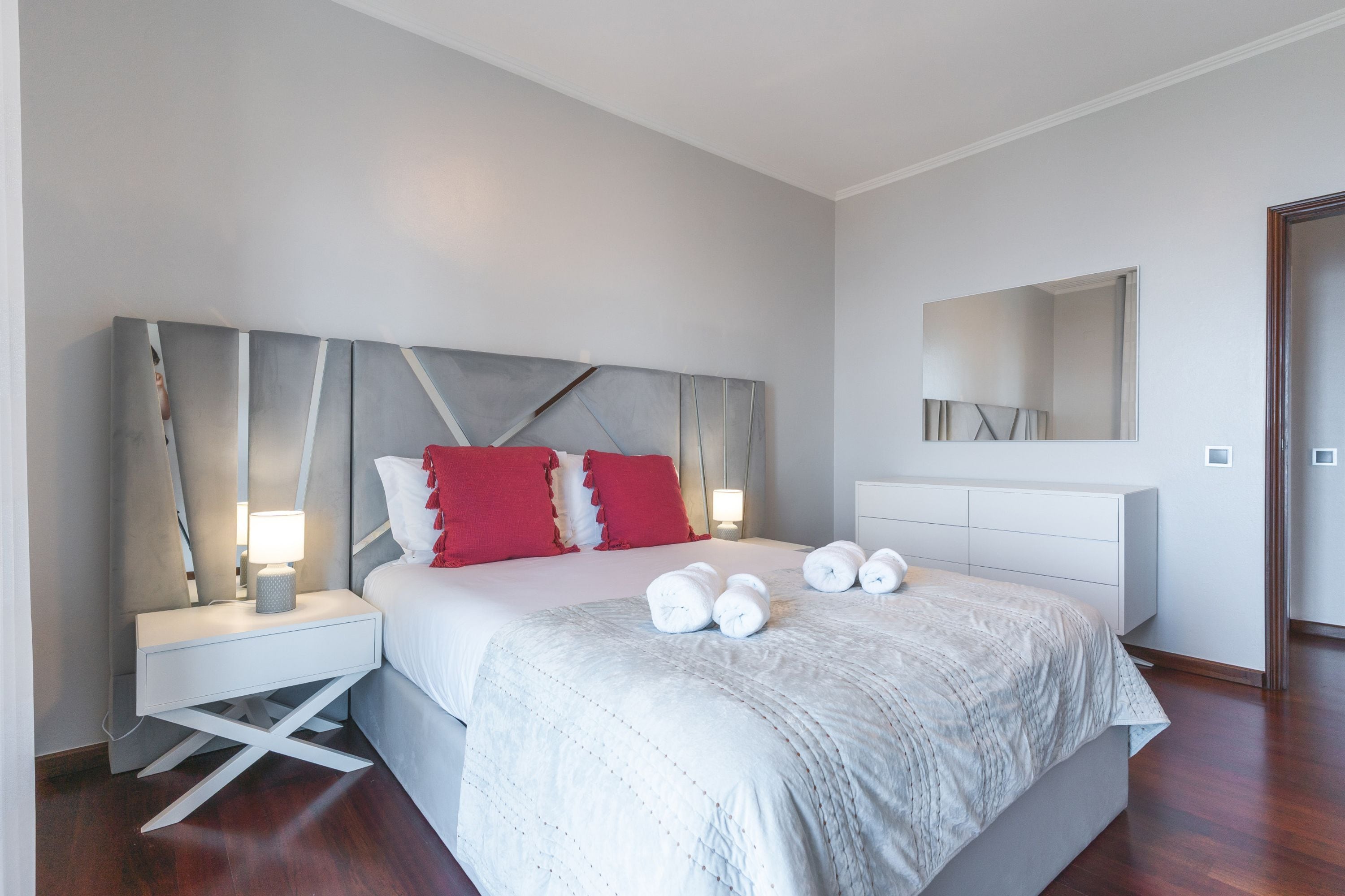 Ein modernes Schlafzimmer mit einem großen Bett, weißen Nachttischen, roten Kissen und einem Spiegel über dem Bett.