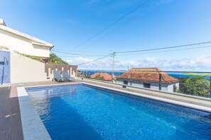 Una piscina con agua azul cristalina, rodeada de una solana con baldosas. Hay sillas de descanso y un pequeño edificio con techo de tejas cerca.