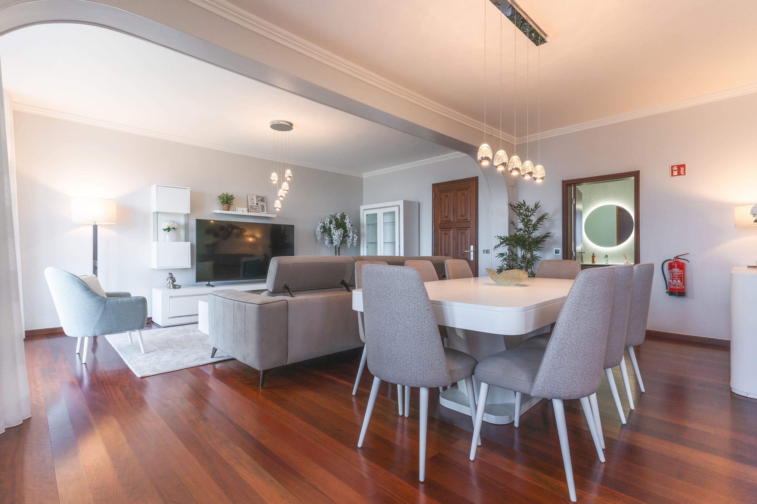 Een moderne woonkamer met een eethoek, houten vloeren, grijze gestoffeerde meubels en hanglampen.