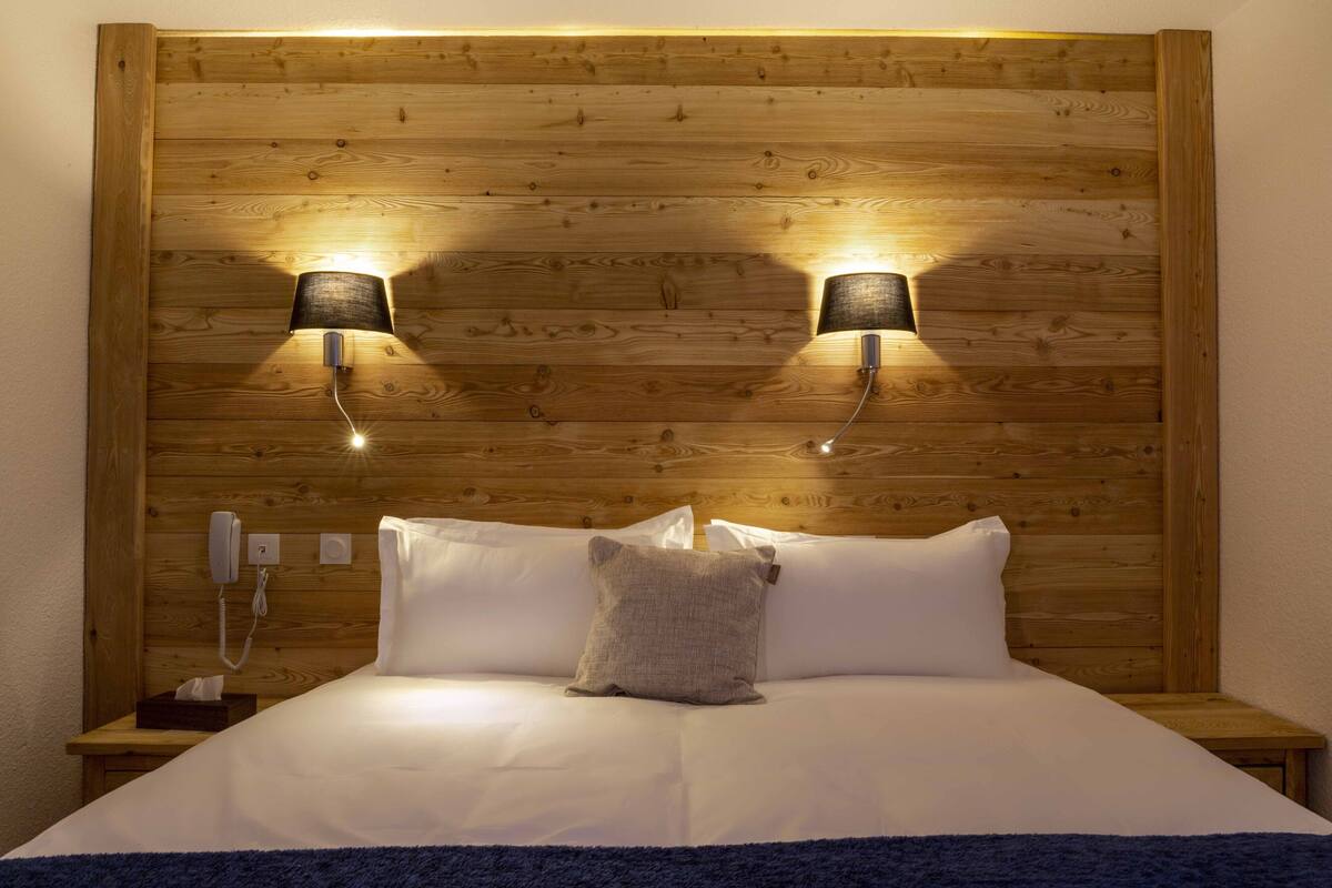 A wooden headboard with two wall-mounted lamps, a bedside phone, and a bed with white linens and a single pillow.