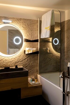 A modern bathroom with a stone wall, a round mirror, a glass-enclosed shower, and a wooden vanity. - Hotel Snow Chill (Risoul)