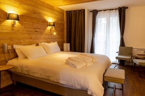 A hotel room with a wooden wall, a bed with white linens, two bedside lamps, a chair, a desk, and a window with curtains. - Hotel Snow Chill (Risoul)