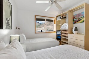 A bedroom with two beds, a window with a mountain view, a ceiling fan, and a wardrobe with shelves and drawers. - Concept 600 Unit 406 (Aspen)