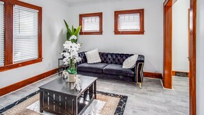 Sala de estar con un sofá azul oscuro, almohadas blancas, una mesa de centro de vidrio y un jarrón con flores blancas. Se observan dos ventanas con marcos de madera y persianas blancas.