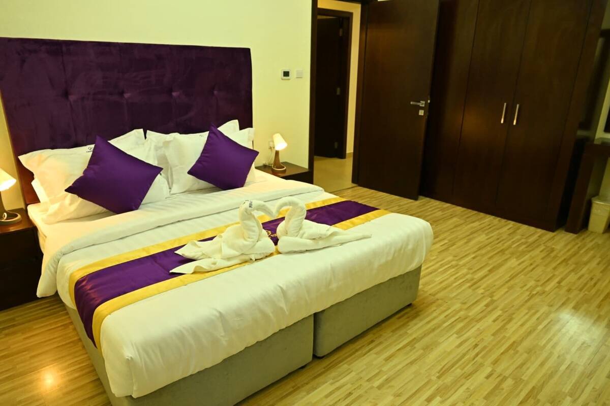 A neatly made bed with purple and white bedding, a pair of white towels arranged in a swan shape, and a wooden floor.