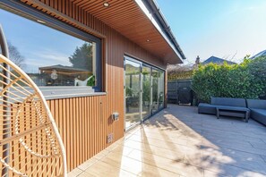 A modern house with a wooden exterior, large sliding glass doors, a patio with a sofa and table, and a wicker chair.