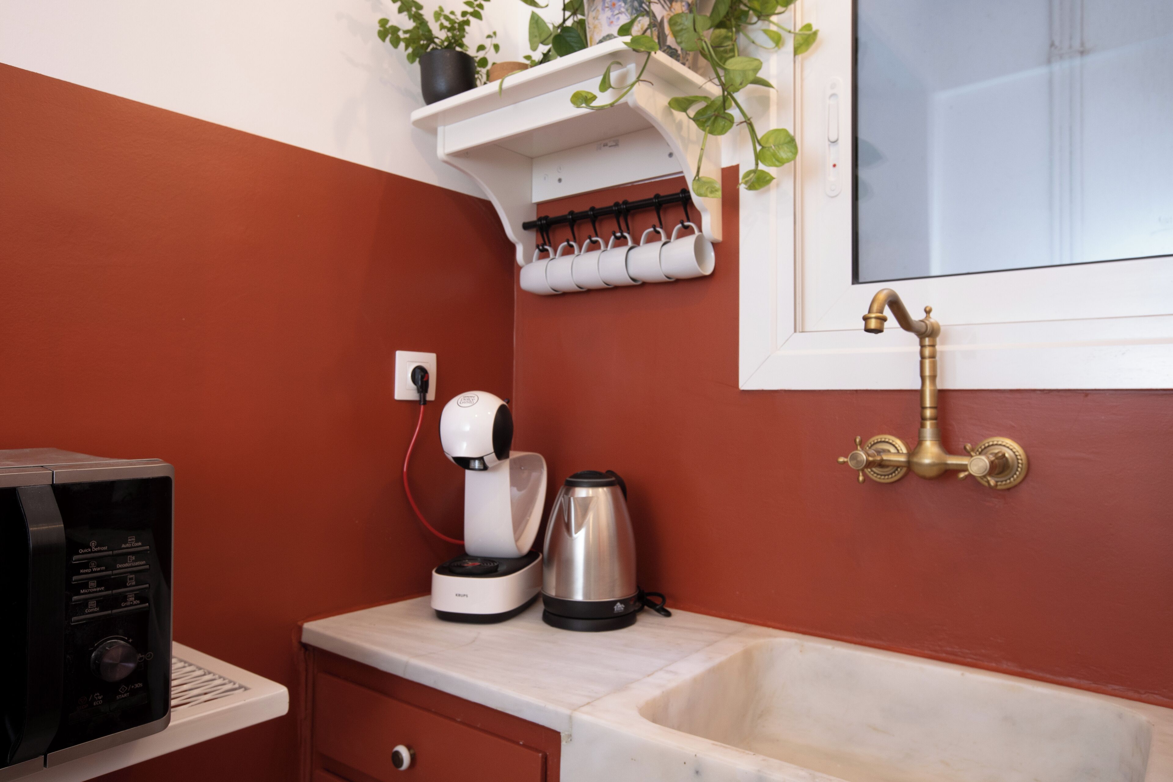 Un baño con pared roja, un lavamanos blanco, un grifo de latón, un estante blanco con una planta y una cafetera.