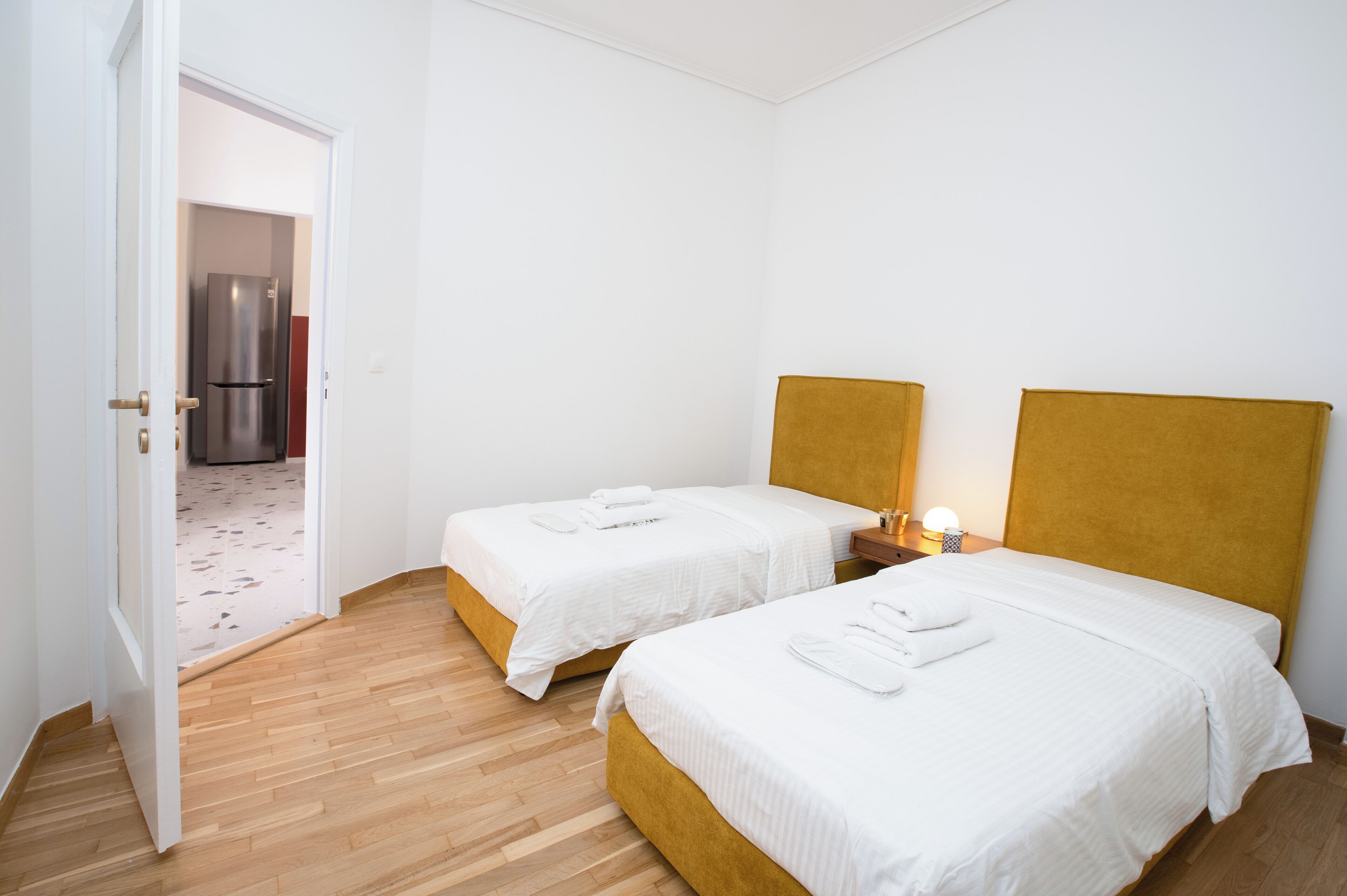 A hotel room with two single beds, wooden flooring, a white door, and a visible kitchen area.