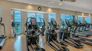 Une salle de sport équipée de plusieurs tapis de course et de plusieurs appareils elliptiques, de grandes fenêtres offrant une vue sur le ciel et les palmiers, et d’un ventilateur de plafond.