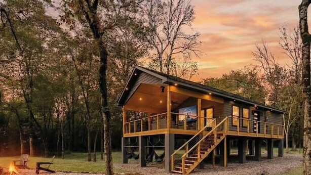 A wooden cabin with a balcony, a staircase, and a fire pit in a forest.