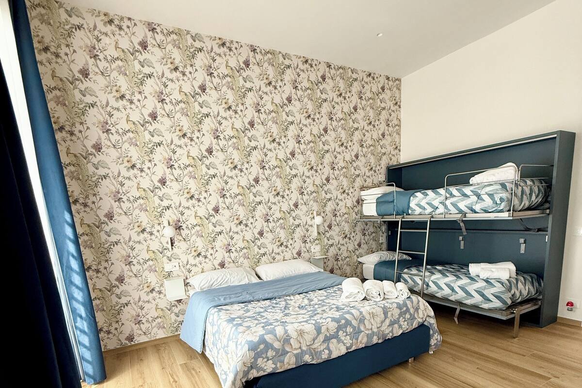 A room with a floral wallpaper, a wooden floor, a bed with a blue floral bedspread, and a bunk bed with patterned bedding.