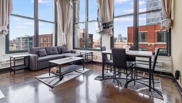 A modern living room with a sofa, coffee table, and dining area. Large windows offer views of the cityscape. - Modern West Loop Gem-City Views & Prime Location 1 (Chicago)