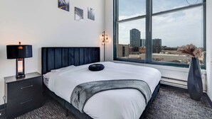 A hotel room with a large window, a bed with white bedding, a black nightstand with a lamp, and artwork on the wall. - Modern West Loop Gem-City Views & Prime Location 1 (Chicago)