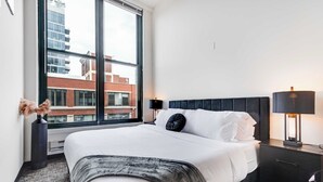 A hotel room with a large window, a bed with white linens, a dark headboard, a bedside table with a lamp, and a decorative vase. - Modern West Loop Gem-City Views & Prime Location 2 (Chicago)