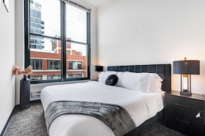 A hotel room with a large window, a bed with white linens, a dark headboard, a bedside table with a lamp, and a decorative vase.