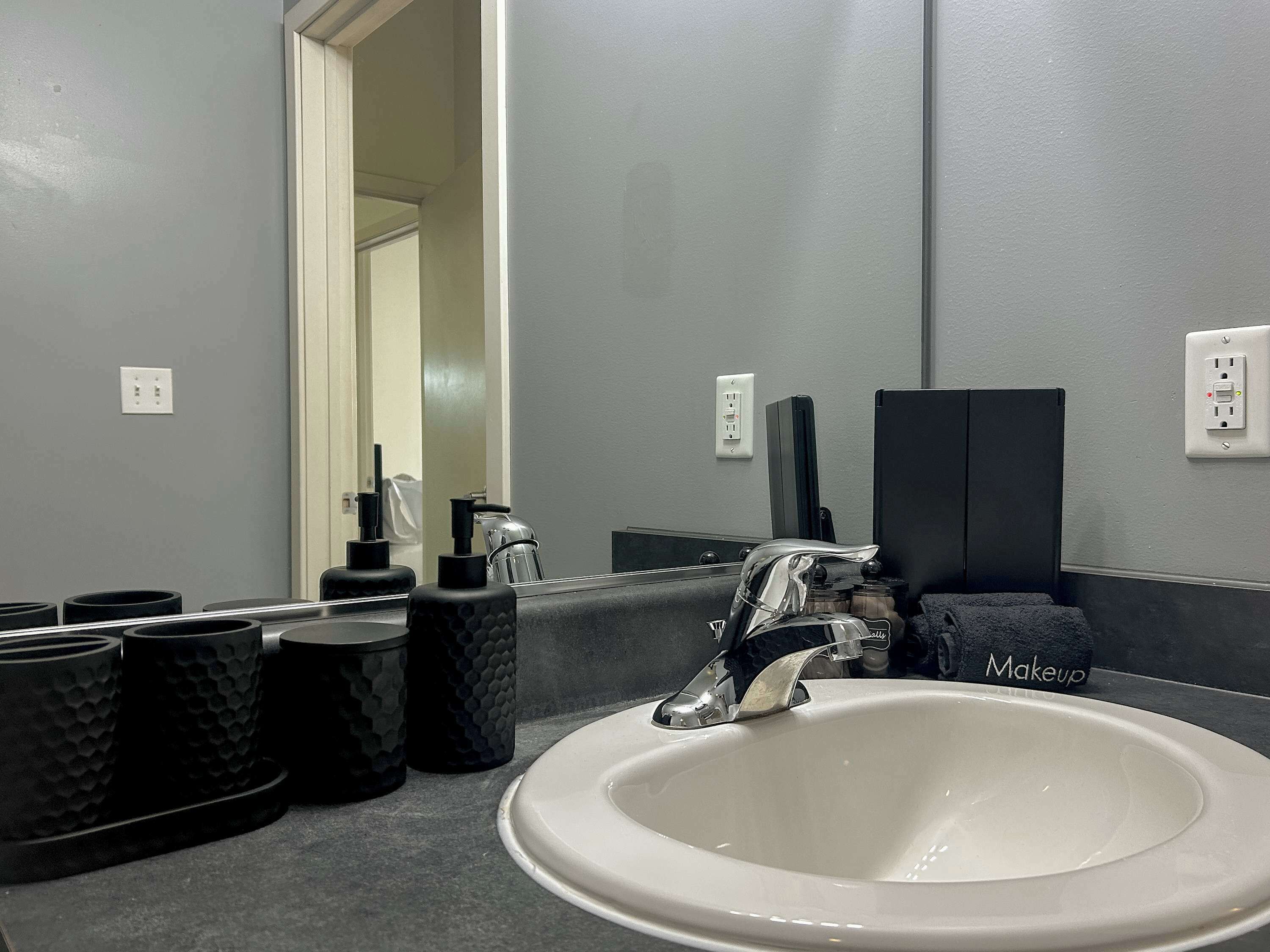 Une salle de bain avec un lavabo blanc, des récipients noirs à texture et un miroir qui reflète une porte.