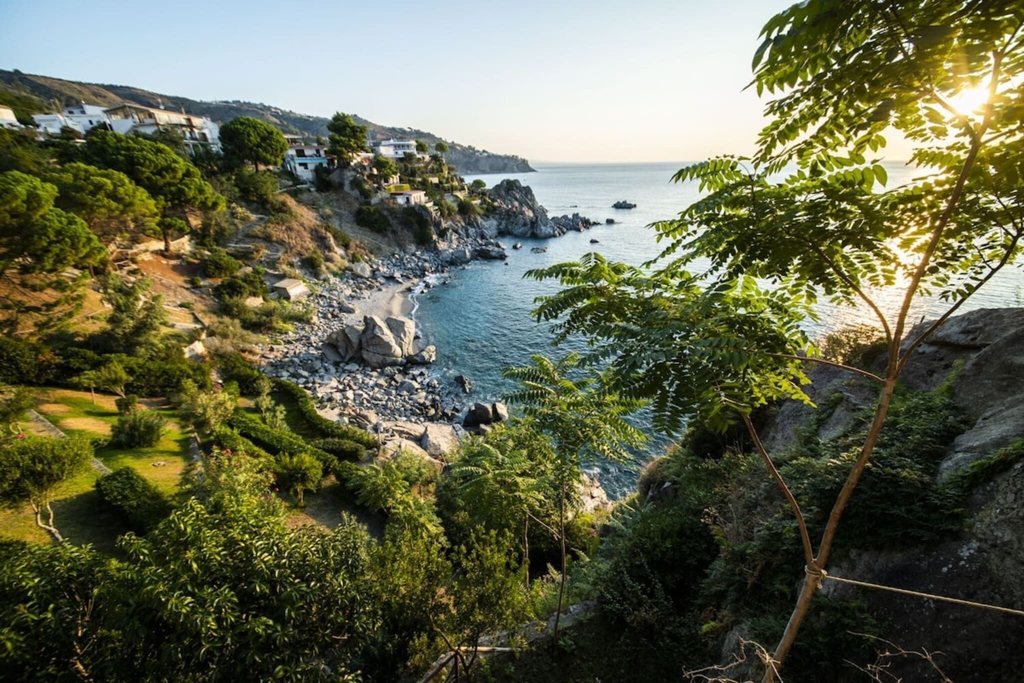 A coastal landscape with rocky terrain, dense greenery, and a view of the sea.