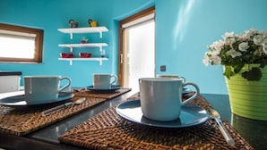 Ein Zimmer mit türkisblauen W änden, einem Fenster und einer Tür. Auf einem Tisch mit einem Löffel stehen zwei weiße Tassen. Eine Zimmerpflanze mit weißen Blüten ist rechts zu sehen.