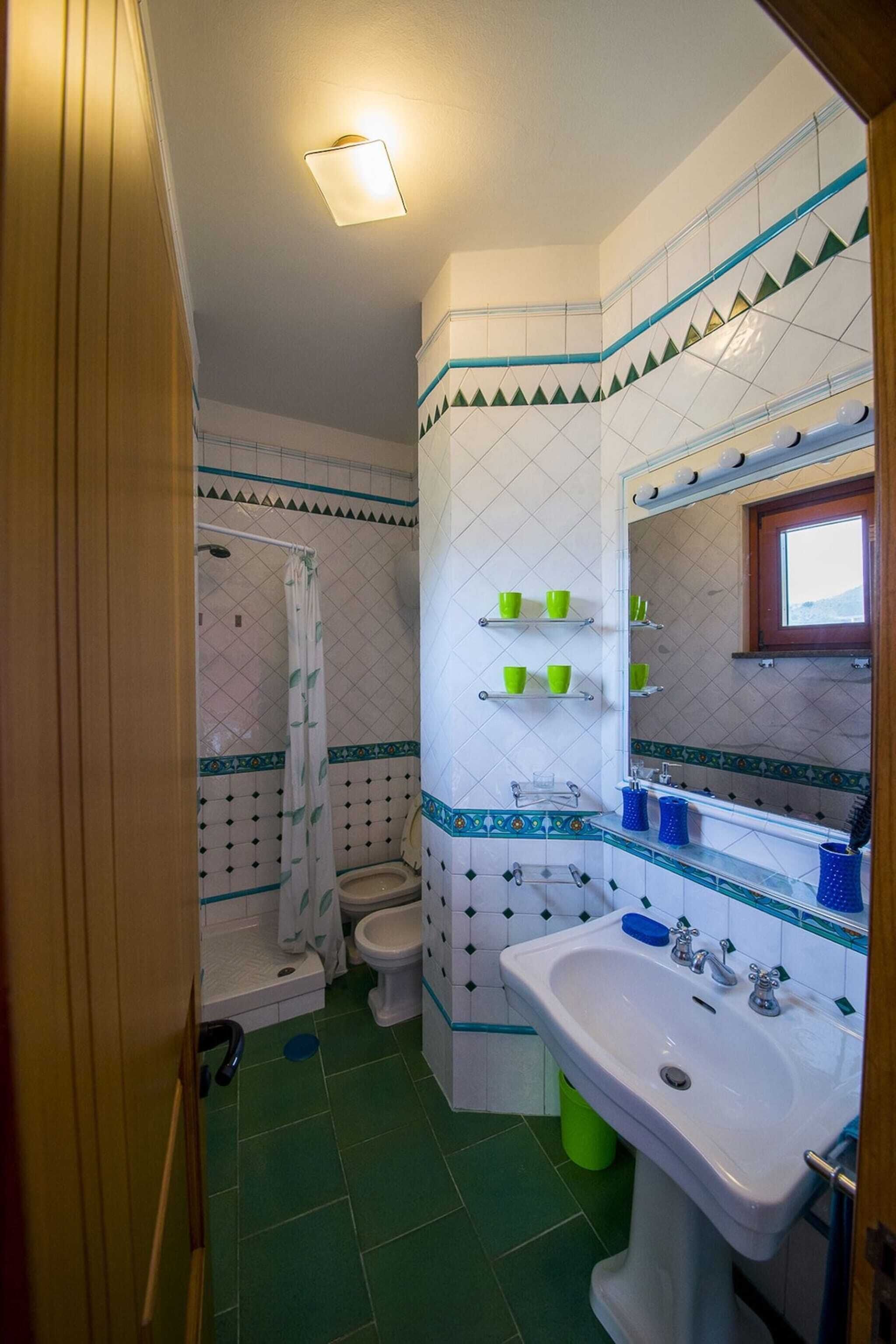Een badkamer met douche, toilet en wastafel, met groene en witte tegels en een spiegel met een raam boven de wastafel.