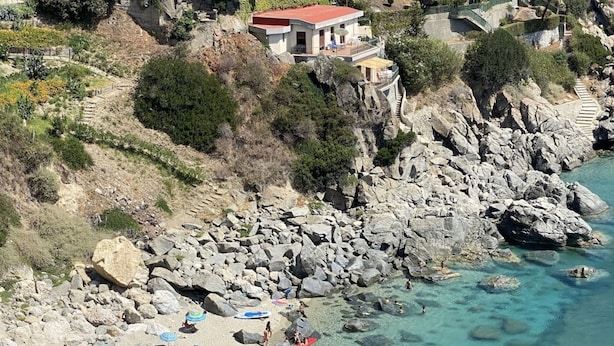 Ein felsiger Strand mit klarem türkisblauem Wasser, Sonnenschirmen und Menschen, die das Meer genießen.