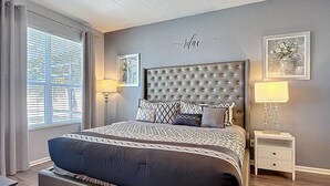 A bedroom with a tufted headboard, patterned bedding, a bedside table with a lamp, and a window with blinds. - My Disney Retreat | Free Resort Access-Disney 9min (Four Corners)