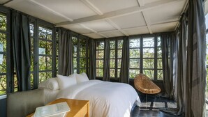 Une pièce avec un grand lit, une table en bois sur laquelle repose un livre, une chaise en osier et de grandes fenêtres avec des rideaux.