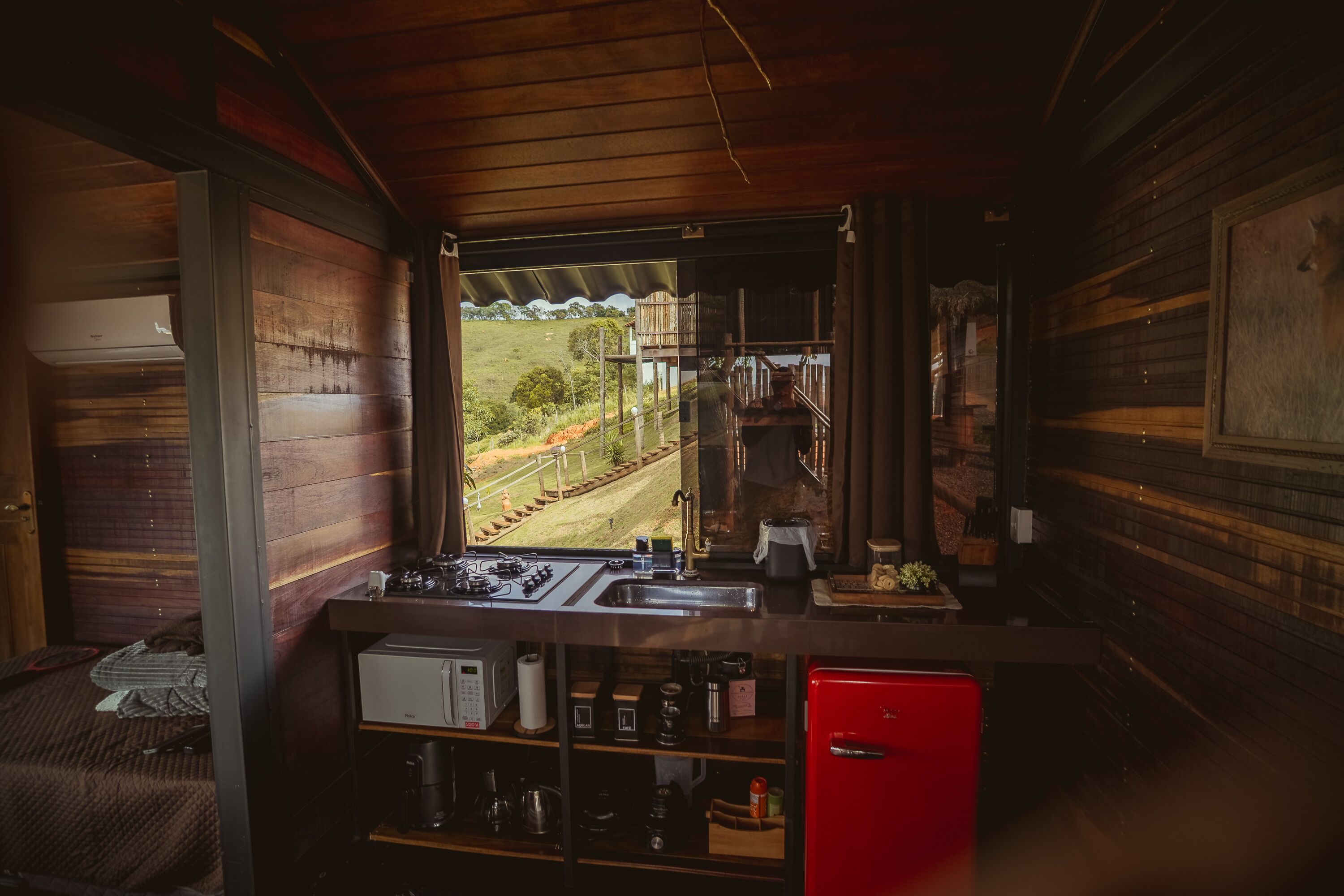 A compact kitchen with a view of a railway track, wooden walls, and a red refrigerator.