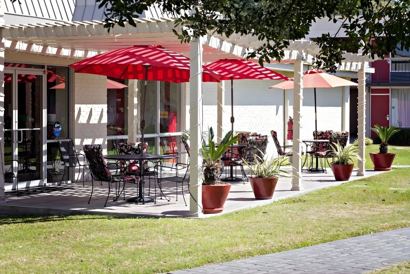 Een buitenterras met tafels en stoelen onder rode parasols, omringd door potplanten.
