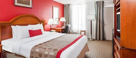 A hotel room with a large bed, a wooden headboard, a desk with a chair, a TV, and a wardrobe.