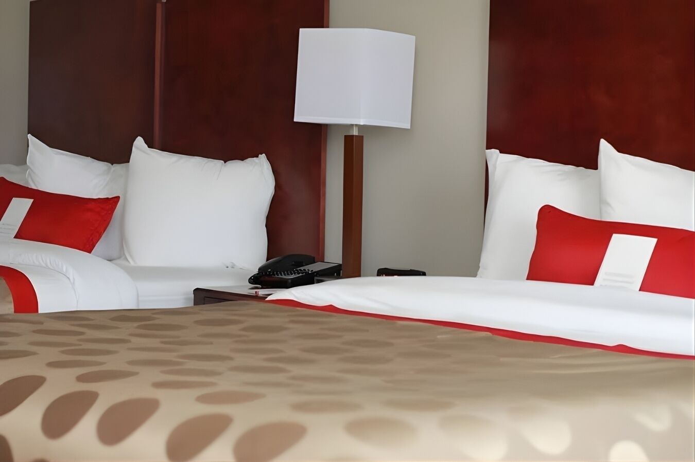 Habitación de hotel con una cama grande, dos almohadas blancas y una roja, una mesita de noche con un teléfono y una lámpara.