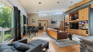 A modern living room with a sofa, a dining area with chairs, a kitchen with wooden cabinets, and a large window offering a view of greenery. - Riverfront City Escape w/Hot Tub, 2 King, Gym, Sleeps 12, Firepit, Kayaks (Winnipeg)